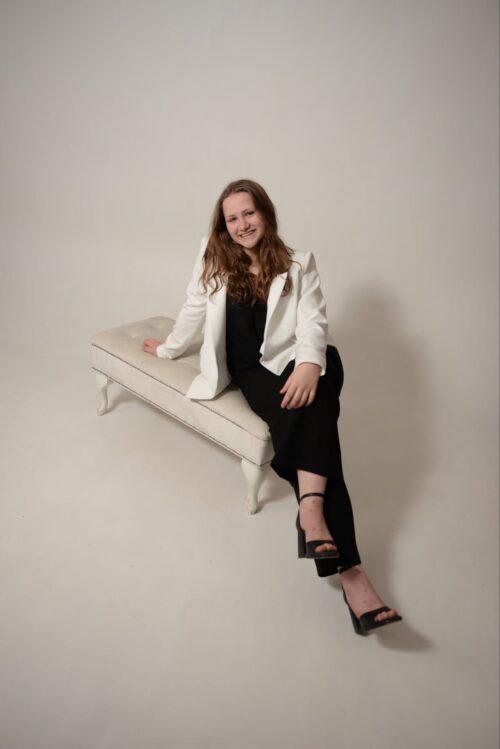Woman in white blazer and black dress sits on a white bench in a studio, smiling at the camera.