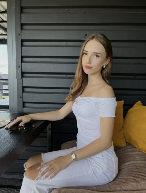 Young woman in a white off-shoulder top sitting at a dark wooden table on a patio, with yellow cushions behind her.