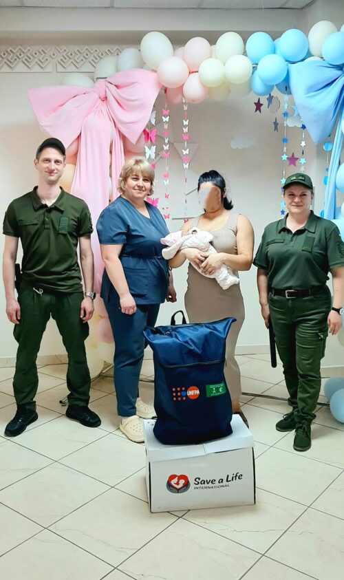 Mother cradling a newborn while two medical staff and two uniformed personnel stand beside her in a balloon‑decorated room, a 'Save a Life' box in front.