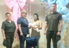 Four adults stand in a room decorated with pastel balloon arches; two wear olive uniforms, two civilians (one woman in blue scrubs) hold a baby doll while a blue gift bag sits on a white box labeled “Save a Life.”