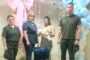 Four adults stand in a room decorated with pastel balloon arches; two wear olive uniforms, two civilians (one woman in blue scrubs) hold a baby doll while a blue gift bag sits on a white box labeled “Save a Life.”