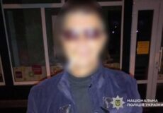 Person in blue police jacket with badge, face blurred, standing outside glass doors; National Police of Ukraine watermark.