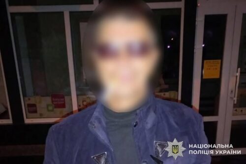 Person in blue police jacket with badge, face blurred, standing outside glass doors; National Police of Ukraine watermark.