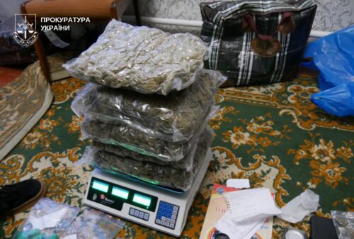 Stack of plastic-wrapped bundles on a digital scale, placed on a patterned carpet; Ukrainian prosecutor logo in the corner.