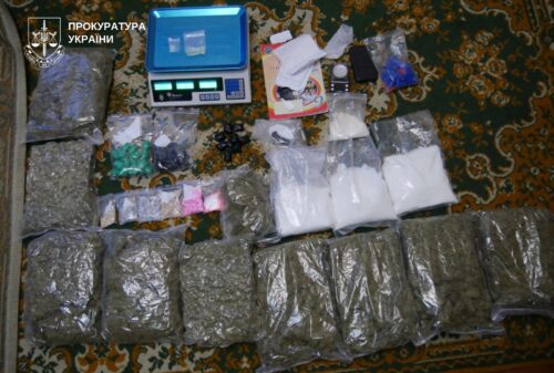 Evidence on an ornate rug: multiple foil-wrapped bundles, small bags, a digital scale, and documents; Ukrainian prosecutors logo in the top-left corner.