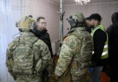 Ukrainian soldiers in camouflage and helmets consult with blurred civilians inside a room.