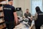 Two law enforcement officers (one with ПРОКУРАТУРА on the back and one with ПОЛІЦІЯ) stand by a cluttered desk as a civilian, face blurred, sits across from them in an office.