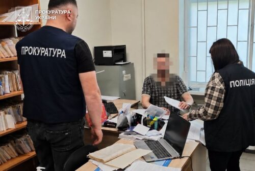 Two law enforcement officers (one with ПРОКУРАТУРА on the back and one with ПОЛІЦІЯ) stand by a cluttered desk as a civilian, face blurred, sits across from them in an office.