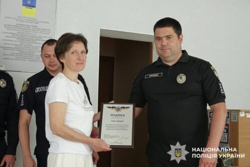 Woman in a white t-shirt presents a framed certificate to a male police officer in a dark uniform; others stand nearby, Ukrainian National Police insignia visible.