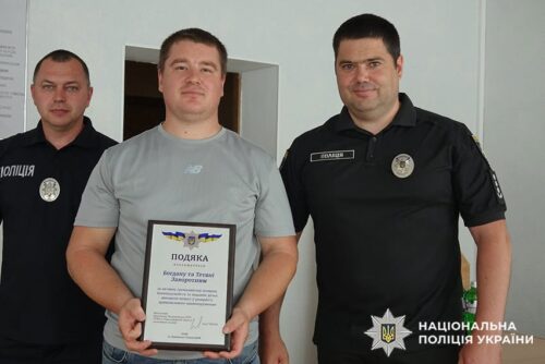 Man in a gray shirt holds a certificate of appreciation as two police officers stand beside him; Ukrainian National Police logo is visible in the corner.