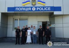 Group of Ukrainian police officers standing with two civilians holding certificates in front of a police station with Ukrainian flags.