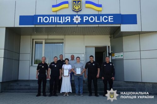 Group of Ukrainian police officers standing with two civilians holding certificates in front of a police station with Ukrainian flags.