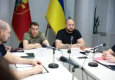 Group of men seated around a white conference table with several microphones and papers, two flags (one blue-yellow Ukrainian flag) behind them, in a formal meeting room.