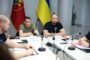 Group of men seated around a white conference table with several microphones and papers, two flags (one blue-yellow Ukrainian flag) behind them, in a formal meeting room.