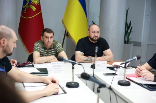 Group of men seated around a white conference table with several microphones and papers, two flags (one blue-yellow Ukrainian flag) behind them, in a formal meeting room.