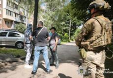 Group of civilians converse on a residential street while a uniformed soldier stands on the right, observing; Ukrainian police emblem shown at bottom right.