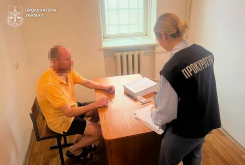 Man in a yellow shirt sits at a desk with papers as a prosecutor in a vest labeled 'ПРОКУРАТУРА' reviews documents in a small office.