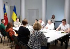 People in a formal meeting around a white conference table; a woman speaks at a podium while flags stand in the background.