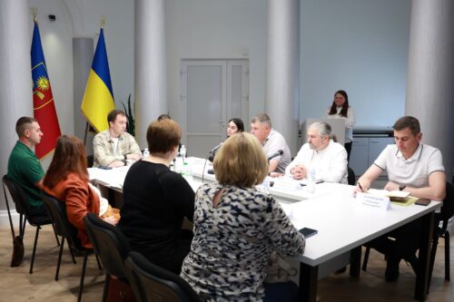 People in a formal meeting around a white conference table; a woman speaks at a podium while flags stand in the background.