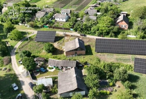 Aerial view of a rural Ukrainian village with wooden houses and large solar panels on rooftops and in nearby fields.