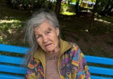 Elderly woman with gray hair sits on a bright blue bench in a park, wearing a multicolored jacket over a brown knit sweater.