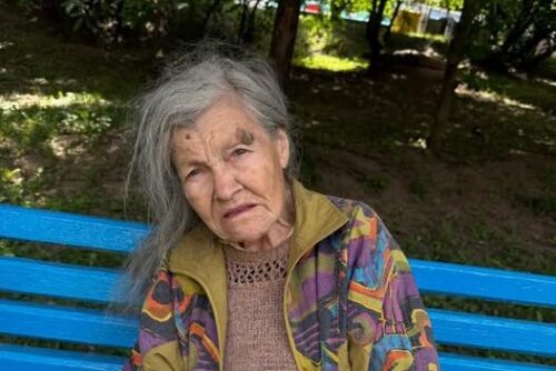 Elderly woman with gray hair sits on a bright blue bench in a park, wearing a multicolored jacket over a brown knit sweater.