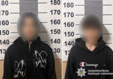 Two people with blurred faces stand before a height-measurement wall in a police booking photo; both wear black hoodies, with police branding visible on the right.