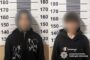 Two people with blurred faces stand before a height-measurement wall in a police booking photo; both wear black hoodies, with police branding visible on the right.