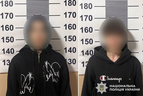 Two people with blurred faces stand before a height-measurement wall in a police booking photo; both wear black hoodies, with police branding visible on the right.