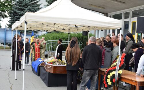 Outdoor funeral under a white canopy with clergy in ceremonial robes beside a closed casket and mourners gathered nearby.