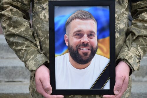 Soldier in camouflage holding a framed portrait of a smiling man with a Ukrainian flag in the background.