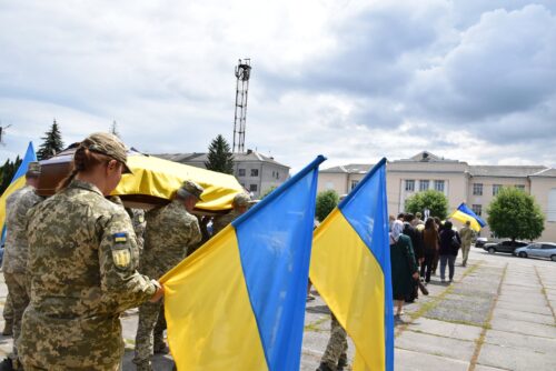 Soldiers in camouflage carry a flag-draped coffin in blue and yellow during a military funeral ceremony.
