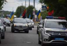 Police SUV leads a street parade, with Ukrainian flags on cars and onlookers nearby.