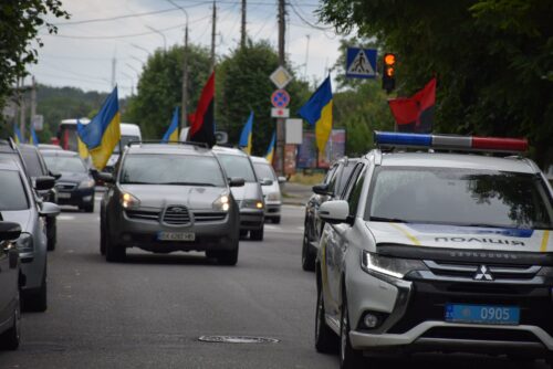 Police SUV leads a street parade, with Ukrainian flags on cars and onlookers nearby.