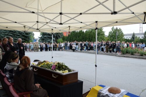 Outdoor funeral under a white canopy; coffin with flowers at the front and a crowd gathered around along the street.