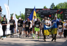 Group of runners crossing a finish-line area, some waving a blue‑yellow Ukrainian flag and smaller banners as spectators look on.