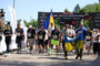 Group of runners crossing a finish-line area, some waving a blue‑yellow Ukrainian flag and smaller banners as spectators look on.
