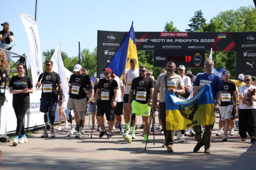 Group of runners crossing a finish-line area, some waving a blue‑yellow Ukrainian flag and smaller banners as spectators look on.