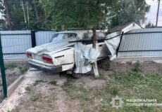 White sedan severely damaged at rear, crushed against a tree beside a metal fence in a yard; police watermark in bottom-right corner.