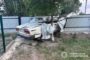 White sedan severely damaged at rear, crushed against a tree beside a metal fence in a yard; police watermark in bottom-right corner.