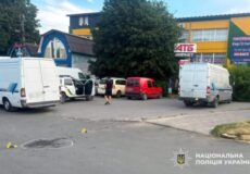 Parking lot outside a yellow storefront area with several white vans and a red van; a few people stand nearby as small yellow evidence markers are on the ground, indicating an ongoing police investigation.
