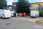 Parking lot outside a yellow storefront area with several white vans and a red van; a few people stand nearby as small yellow evidence markers are on the ground, indicating an ongoing police investigation.