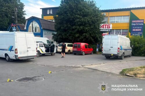 Parking lot outside a yellow storefront area with several white vans and a red van; a few people stand nearby as small yellow evidence markers are on the ground, indicating an ongoing police investigation.