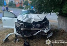 White car with severe front-end damage after a crash into a tree; driver's door open and police presence in the background.