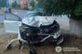 White car with severe front-end damage after a crash into a tree; driver's door open and police presence in the background.