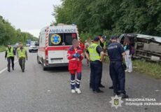 Emergency responders, an ambulance, and police at a highway crash; a vehicle lies on its side by a grassy verge behind them.