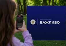 Person using a smartphone to photograph a blue sign with a police emblem and the word 'ВАЖЛИВО' outdoors in a park.