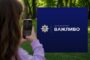 Person using a smartphone to photograph a blue sign with a police emblem and the word 'ВАЖЛИВО' outdoors in a park.