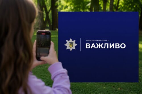 Person using a smartphone to photograph a blue sign with a police emblem and the word 'ВАЖЛИВО' outdoors in a park.