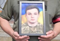 Person in a green military uniform holding a framed photo of a young man in camouflage.